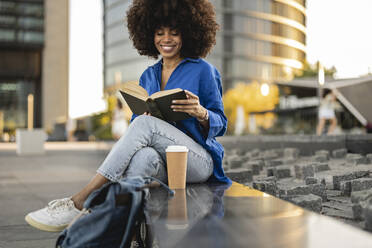 Smiling Afro woman reading book sitting by disposable coffee cup on wall - JCCMF07382