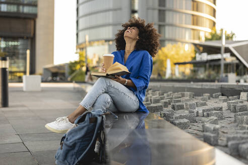 Fröhliche Afro-Frau mit Buch und Einwegbecher an der Wand sitzend - JCCMF07383