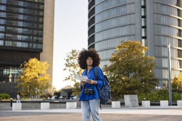 Woman with Afro hairstyle holding map in front of buildings - JCCMF07396