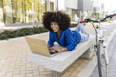 Happy woman using laptop lying on concrete bench by bicycle - JCCMF07398