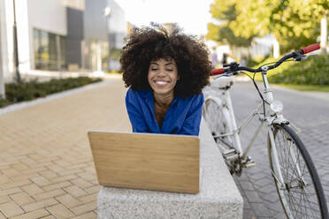 Lächelnde Afro-Frau mit Laptop auf einer Betonbank neben einem Fahrrad liegend - JCCMF07399