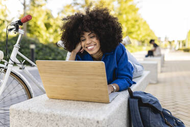 Glückliche Afro-Frau schaut auf einen Laptop, der auf einer Betonbank liegt - JCCMF07400