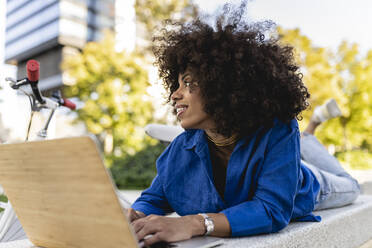 Smiling Afro woman with laptop lying on concrete bench - JCCMF07401
