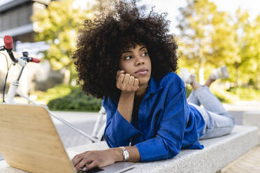 Thoughtful woman with laptop lying on concrete bench - JCCMF07402