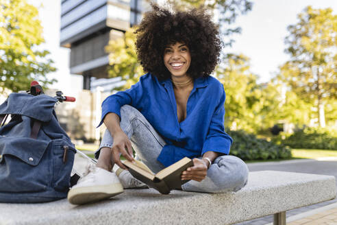Glückliche Afro-Frau mit Buch auf einer Betonbank sitzend - JCCMF07405