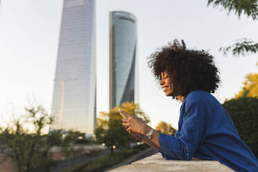 Lächelnde Frau mit Afrofrisur, die ein Smartphone benutzt und sich an eine Wand lehnt - JCCMF07417