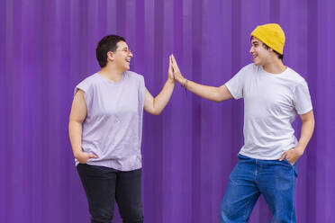 Happy non-binary couple giving high-five in front of corrugated wall - EGCF00044