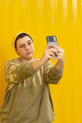 Nicht-binäre Person, die ein Selfie mit ihrem Smartphone vor einer gelben Wellblechwand macht - EGCF00049