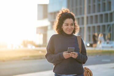 Smiling businesswoman with smart phone standing at footpath on sunny day - JOSEF13352