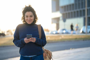 Smiling businesswoman with smart phone standing on footpath - JOSEF13354