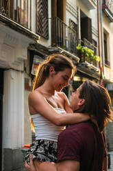 Side view of smiling cheerful couple in casual wear standing on street and looking at each other while boyfriend picking up young woman in arms during spending time together - ADSF39234