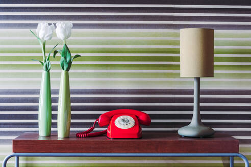Blumen in grünen Vasen und Retro-Telefon neben Lampe in Reihe auf Holztisch gegen Retro-Tapete platziert - ADSF39236