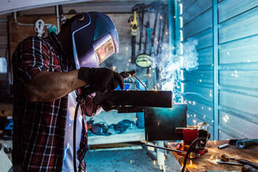 Middle aged casual carpenter in helmet and gloves heating metal part with gas torch working in garage - ADSF39247