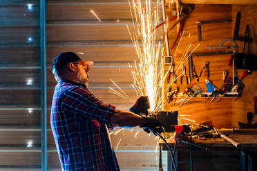 Seitenansicht eines reifen männlichen Arbeiters, der einen Winkelschleifer benutzt, während er ein Metallteil mit glühenden Funken in einer Garage bearbeitet - ADSF39248