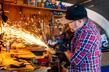 Seitenansicht eines reifen männlichen Arbeiters, der einen Winkelschleifer benutzt, während er ein Metallteil mit glühenden Funken in einer Garage bearbeitet - ADSF39249