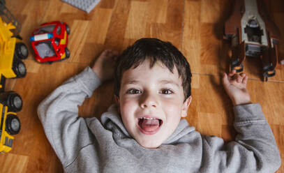 Von oben von einem fröhlichen kleinen Jungen, der auf dem Parkettboden liegt und in die Kamera schaut, während er zu Hause mit Spielzeugautos spielt - ADSF39295