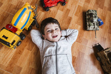 Von oben von einem fröhlichen kleinen Jungen, der auf dem Parkettboden liegt und in die Kamera schaut, während er zu Hause mit Spielzeugautos spielt - ADSF39296