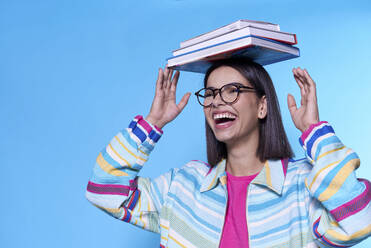 Happy young woman wearing colorful zipper balancing books on head against blue background - LCZF00002