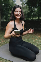 Lächelnder Yogalehrer mit Mobiltelefon auf einer Übungsmatte im Park sitzend - MRRF02469
