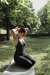 Yogalehrer, der auf einer Übungsmatte im Park sitzend die Haare zusammenbindet - MRRF02476