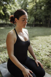 Yoga teacher with eyes closed on exercise mat in park - MRRF02487