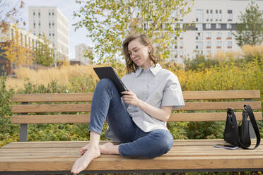 Junge Frau sitzt auf einer Bank und benutzt einen Tablet-PC im Park - LESF00321
