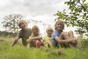 Smiling brothers and sister with blond hairs sitting on grass - NJAF00006