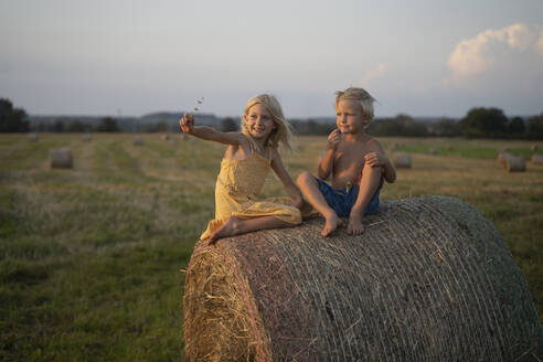 Lächelndes Mädchen mit ausgestrecktem Arm, das neben seinem Bruder auf einem Heuhaufen sitzt - NJAF00014