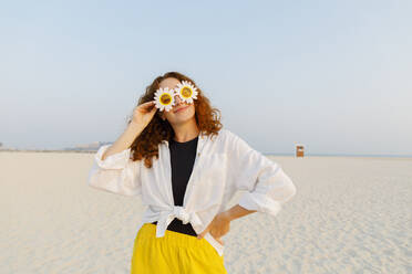 Lächelnde Frau mit Sonnenblumen-Sonnenbrille, stehend mit Hand auf der Hüfte am Strand - TYF00430