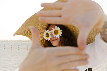 Lächelnde Frau mit Sonnenblumen-Sonnenbrille und übergroßem Strohhut gestikuliert am Strand - TYF00432