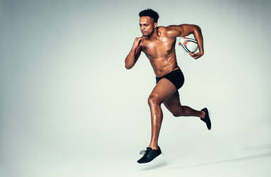 Full length shot of muscular young man running with a rugby ball on grey background. African male rugby player with muscular build. - JLPSF00004