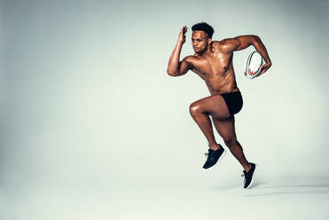 Full length shot of young rugby player training. Muscular male model running with rugby ball on grey background. - JLPSF00005