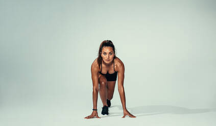 Totale Aufnahme einer selbstbewussten Athletin in Startposition, bereit für einen Lauf. Hispanische junge Frau, die einen Sprint beginnt, vor grauem Hintergrund. - JLPSF00039