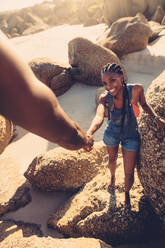 Junge Frau, die mit Unterstützung ihres Freundes durch die Felsen läuft. POV-Aufnahme eines Mannes, der seiner Freundin beim Klettern auf einem Felsen am Strand hilft. - JLPSF00045