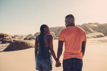 Ein afroamerikanisches Paar, das sich an den Händen hält und am Strand spazieren geht. Ein verliebter Mann und eine verliebte Frau spazieren an einem Sommertag am Meer entlang - JLPSF00046