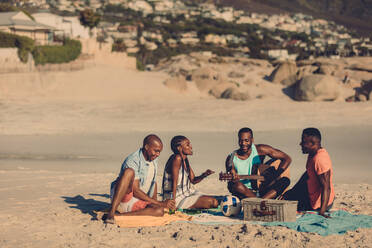 Gruppe von Freunden mit Gitarre am Strand sitzend. Afrikanische Menschen genießen ein Picknick am Meer. - JLPSF00050