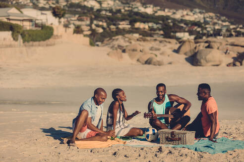 Gruppe von Freunden mit Gitarre am Strand sitzend. Afrikanische Menschen genießen ein Picknick am Meer. - JLPSF00050