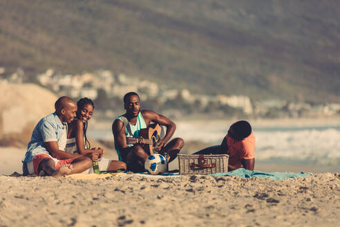 Afroamerikanischer junger Mann mit Gitarre, der ein Lied für seine Freunde singt. Gruppe von Freunden im Urlaub, die sich am Strand entspannen. - JLPSF00051