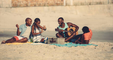 Eine Gruppe junger Leute hängt am Strand ab. Ein junger afroamerikanischer Mann spielt Gitarre für seine Freunde. - JLPSF00052