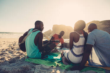 Afroamerikanischer Mann spielt Gitarre am Strand mit Freunden, die um ihn herum sitzen. Menschen am Strand genießen ihren Urlaub. - JLPSF00057