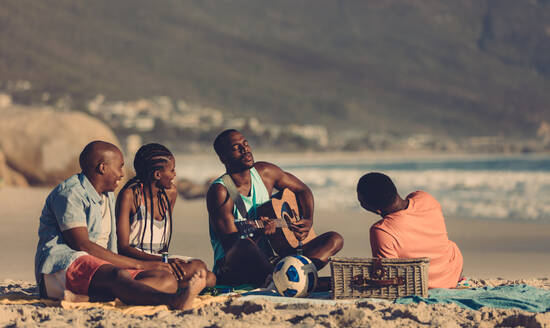 Afrikanischer Mann spielt Gitarre für Freunde am Strand. Gruppe von Freunden beim Picknick am Meer. - JLPSF00061