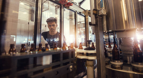 Ein Fabrikarbeiter schaut auf ein Förderband, auf dem sich Bierflaschen bewegen. Ein Brauer überwacht den Prozess der Bierabfüllung in der Produktionsstätte. - JLPSF00064