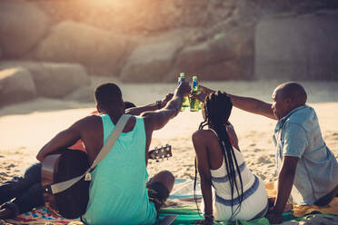 Junge Leute stoßen mit Bierflaschen an, während sie am Strand sitzen. Eine Gruppe von Freunden, die zusammen etwas trinken. - JLPSF00065
