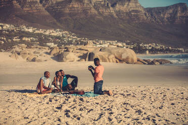 Afrikanischer junger Mann fotografiert seine Freunde mit dem Handy am Strand. Gut aussehender Mann fotografiert seine Freunde mit dem Smartphone. - JLPSF00067