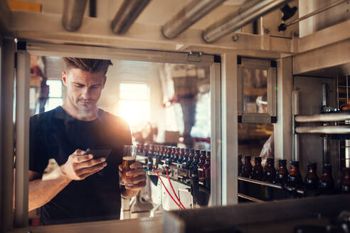 Young man with a glass of beer at brewery plant using mobile phone. Beer factory owner at bottling machine reading text message on his smart phone. - JLPSF00072