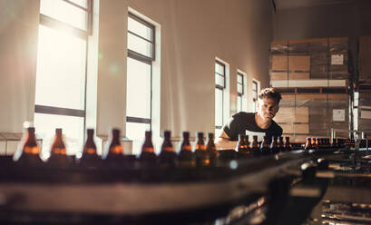 Ein Brauer schaut auf ein Förderband mit Bierflaschen. Ein junger Mann überwacht den Herstellungsprozess von Bier in einer Brauerei. - JLPSF00087