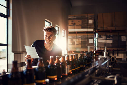 Junger Mann, der den Prozess der Bierherstellung in einer Brauereifabrik überwacht. Fabrikarbeiter, der den Prozess am Fließband in der Brauerei kontrolliert. - JLPSF00088