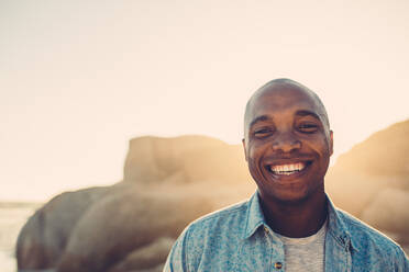 Porträt eines afrikanischen Mannes am Strand, der lächelnd in die Kamera schaut, an einem sonnigen Tag. - JLPSF00092