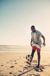 Junger Mann kickt Fußball am Meer. Schwarzer Mann spielt mit Fußball am Strand. - JLPSF00098