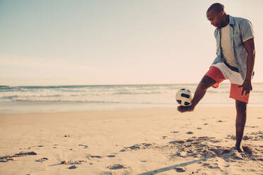 Afroamerikanischer Mann spielt mit Fußball am Strand. Schwarzer Mann balanciert einen Fußball auf seinen Füßen am Meeresufer. - JLPSF00099